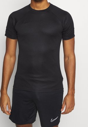 Mann trägt schwarzes, kurzärmeliges Nike-Sportshirt und schwarze Nike-Shorts und steht vor einfarbigem Hintergrund.
