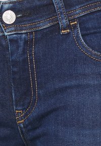 Jeans de mezclilla azul oscuro con costuras naranjas en contraste, que cuentan con un bolsillo frontal y un cierre de botón metálico. Textura suave y diseño ajustado.