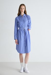 Blåstripet skjortekjole med knapplukking, krage, belte i livet og lange ermer. Modellen har på seg hvite joggesko og ankelsokker.