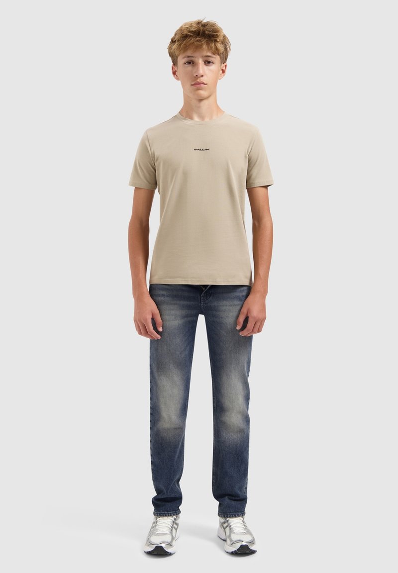 Tiener in een beige T-shirt met korte mouwen, vervaagde blauwe jeans en witte sneakers, staande tegen een effen lichtgrijze achtergrond.