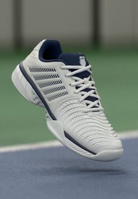 Adidașii albi cu un toc texturat, accente albastre, design cu șireturi și o parte ventilată cu dungi diagonale pentru respirabilitate.