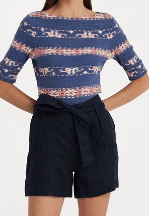 Kobieta nosząca dopasowany top w niebiesko-różowy wzór z rękawami do połowy przedramienia oraz czarne, wysokie szorty zawiązane materiałowym paskiem.