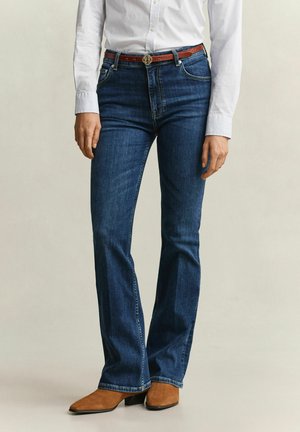 Personne portant un jean bootcut bleu foncé, une ceinture marron, une chemise blanche à boutons et des bottines en daim marron, debout devant un fond uni.