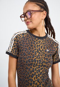 Camiseta marrón con estampado de leopardo, con cuello y mangas negras. Presenta rayas blancas en los hombros y un pequeño logo de la marca en el pecho.