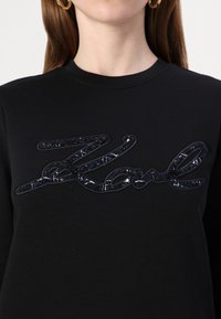 Felpa nera con logo ricamato cursivo lucido in blu scuro. Il design presenta una finitura testurizzata sul logo.