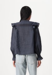 Navy blue blouse with puffed sleeves, lace detailing, and a high neckline. Smooth texture and cropped fit paired with light blue denim jeans.