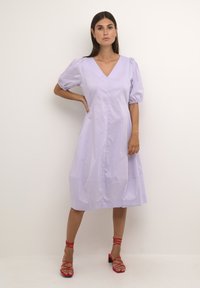 Šviesi levandų spalvos vidutinio ilgio suknelė su pūstomis rankovėmis, V formos iškirpte ir tiesiu siluetu, derinama su raudonais diržiniais sandalais. Lygus audinio paviršius.