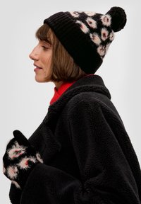 Zwarte gebreide muts met pompon en wit bloemenpatroon; bijpassende handschoenen hebben een vergelijkbaar ontwerp. Model draagt een zwarte fleece jas en een rode shirt.