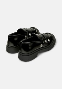 Svarta, blanka slip-on-skor med tjock sula och dekorativa silverknappar, vy från baksidan som visar skoens design och stil.