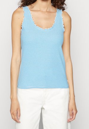 Kvinde iført en blå og hvid stribet ærmeløs bluse med bølgede kanter, parret med hvide bukser, mod en ensfarvet baggrund.