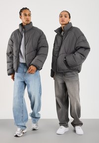 Zwei Personen tragen übergroße, graue Pufferjacken mit hohen Kragen, dazu abgerissene, locker sitzende Jeans und weiße Turnschuhe.