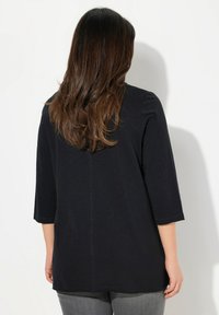 Musta kolmene neljännes hihainen paita, jossa on rento istuvuus, sileä tekstuuri ja hyvin hillitty sauman yksityiskohta keskellä, yhdessä harmaiden housujen kanssa.