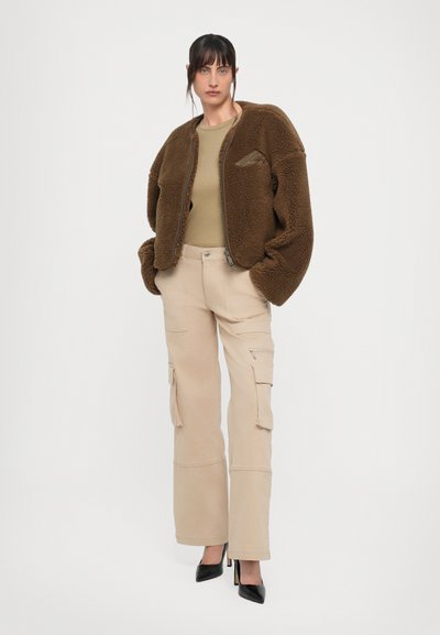 Braune Fleecejacke aus strukturiertem Material, olivgrünes Oberteil, beigefarbene Cargo-Hose mit Taschen und schwarze, spitz zulaufende High Heels.