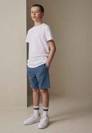 Dječak stoji u zatvorenom prostoru, nosi bijelu majicu, plave kratke hlače, bijele tenisice i bijele čarape s crnim prugama, ruke u džepovima.