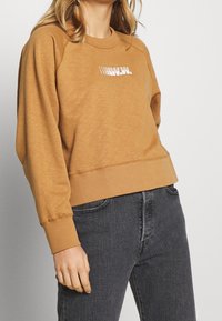 Brun sweatshirt med en ribbad rundhalsringning, något kortad passform och vit grafisk text på framsidan. Bärs med svarta jeans.