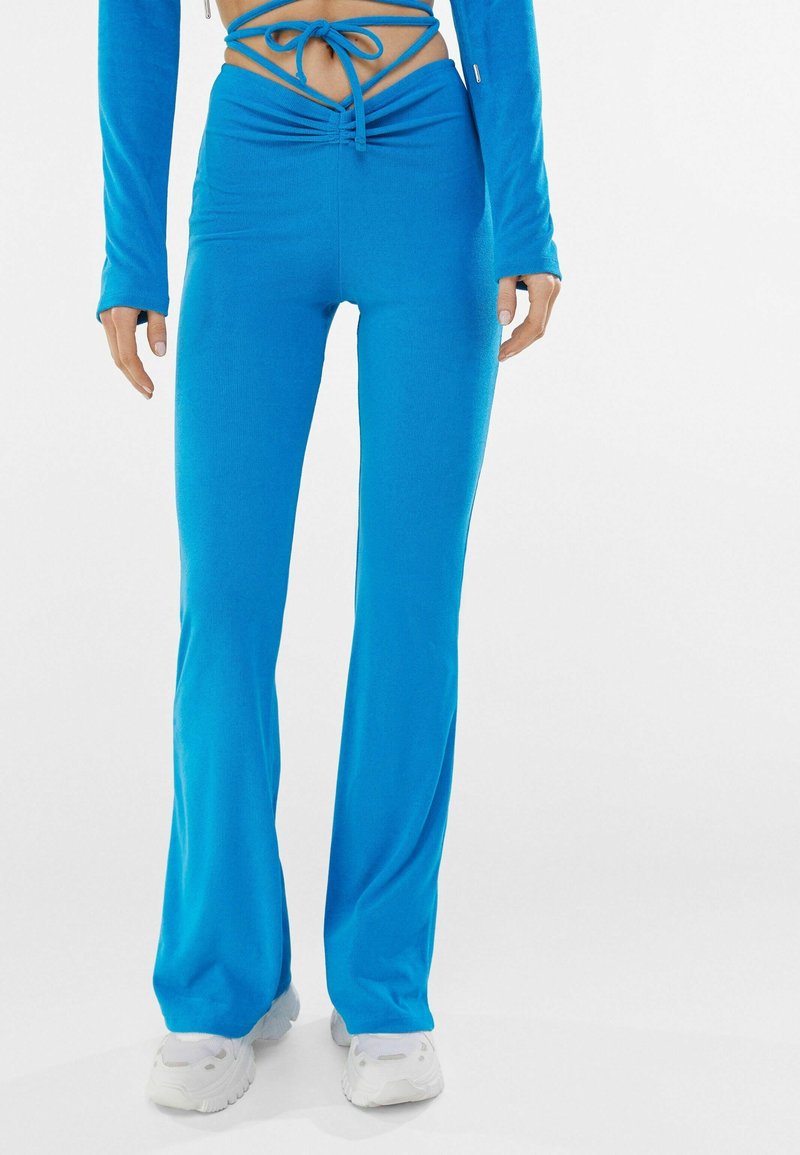 Blue stretch fabric flared pants with a gathered waistband, featuring a drawstring tie and a smooth texture. Paired with white sneakers.