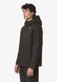 Donkerbruine capuchonjas met hoge kraag, ritssluiting aan de voorkant en steekzakken aan de zijkant. Gladde textuur met een subtiel logo op de borst.