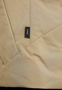 Beżowa tkanina o teksturowanej powierzchni z diagonalnymi wzorami, zawierająca czarną metkę z napisem "CHASIN'". Projekt jest kątowy i warstwowy.