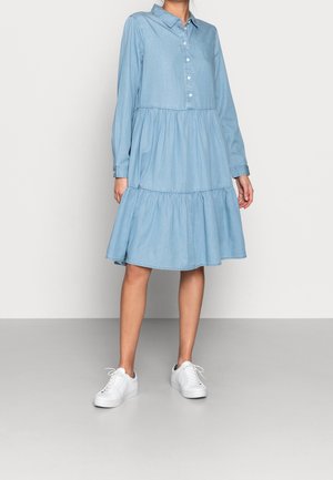 Kvinde iført en lyseblå langærmet kjole med lag og knapper foran samt hvide snørelignende sneakers, stående på en grå gulv.