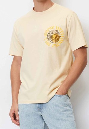 Crèmekleurig T-shirt met een grafische print in gele en bruine tekst met de tekst "Groei met de Stroming," met korte mouwen en een ontspannen pasvorm.