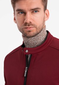 Burgunderfärgad zip-jacka i texturerat tyg, med ståkrage och en ribbad, mönstrad polokrage under. Dragkedja med metalliska dragflik.