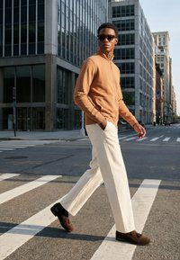 Homme portant une veste marron, un pantalon blanc et des lunettes de soleil marchant sur un passage piéton en ville avec de grands bâtiments en verre en arrière-plan.