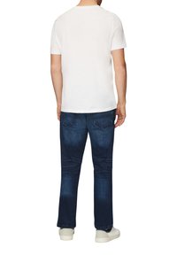 T-shirt en coton blanc à manches courtes, associé à un jean en denim bleu foncé. Le jean présente un design délavé et une couture typique des poches arrière.