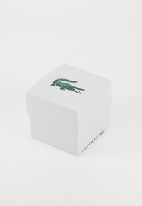 Hvit teksturert pappeske med en grønn krokodillelogo på toppen og "LACOSTE" trykket på siden. Kvadratisk form med lokk.