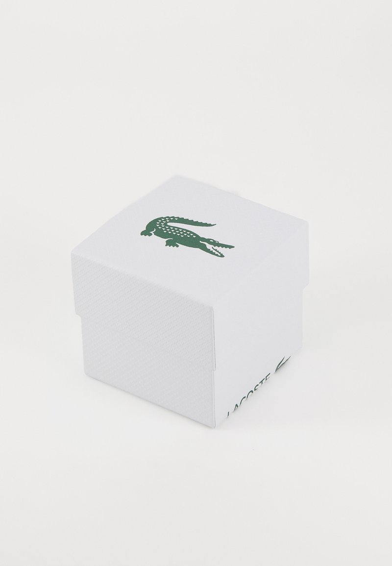 Caja de cartón texturizada blanca con un logo de cocodrilo verde en la parte superior y "LACOSTE" impreso en el lateral. Tiene forma cuadrada y una tapa.