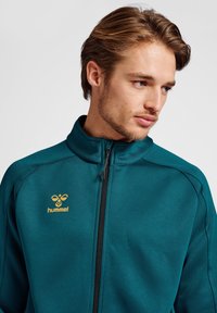 Teal zipjack met hoge kraag, voorzien van een zwarte rits, gestructureerde stof en een gouden "hummel" logo op de borst.