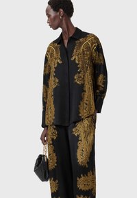 Black satin shirt and pants set with intricate golden paisley patterns. The model holds a small black handbag with gold hardware accents.