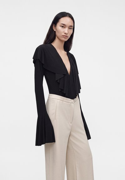 Femme portant un haut noir à manches longues avec un profond décolleté en V et des détails à volants, associé à un pantalon beige large taille haute, debout devant un fond blanc.