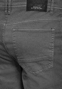 Szare dżinsy mają teksturowane wykończenie z widocznym szwem i tylną kieszenią. Na pasku wszyta jest czarna metka.