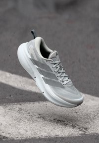 Ανοιχτό γκρι παπούτσι για τρέξιμο Adidas με λευκή σόλα και τρεις ρίγες, αιωρούμενο πάνω από την άσφαλτο με διασκορπισμένα σωματίδια βρωμιάς.