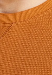 Tejido de algodón naranja con un punto texturizado. En la imagen de cerca se observan el cuello acanalado y los detalles de costura a lo largo de los bordes.