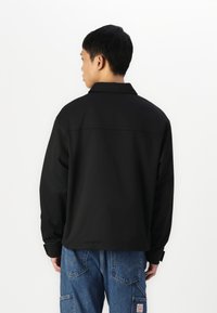 Veste noire oversized avec col, manches longues et coupe droite. Présente une texture lisse et un détail de dos en yoke rectangulaire.