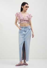 Donna che indossa occhiali da sole beige, top floreale corto con maniche a volant, gonna di jeans alta in vita di un blu chiaro con spacco frontale e sandali viola con lacci.