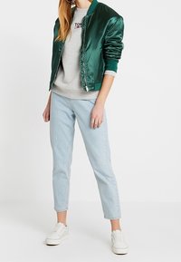 Person wearing a dark green satin bomber jacket, gray sweatshirt, light blue jeans, and white sneakers standing on a white background.