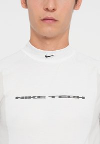 White long-sleeve athletic shirt with a high collar, featuring a textured fabric and a prominent black "NIKE TECH" logo on the front.