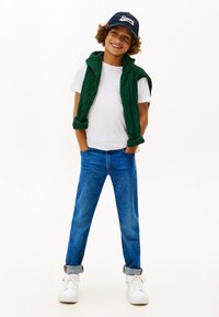 Lächendes Kind mit navyblauer Mütze, weißem T-Shirt, grünem Pullover über den Schultern, hochgekrempelten blauen Jeans und weißen Turnschuhen, steht mit den Händen in den Taschen.