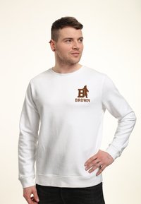 Sweat-shirt blanc avec un col rond. Présente un logo marron avec un ours et le texte "BROWN" sur la poitrine gauche. Design simple, manches et ourlet côtelés.