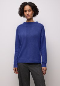 Blauer Strickpullover mit hohem Kragen, strukturierten Einsätzen und langen Ärmeln, kombiniert mit dunkler, strukturierter Hose.