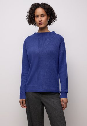 Blauer Strickpullover mit hohem Kragen, strukturierten Einsätzen und langen Ärmeln, kombiniert mit dunkler, strukturierter Hose.