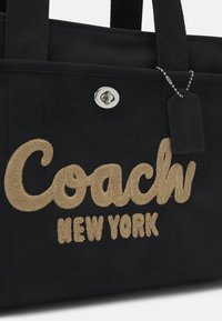 Juodas audinės prekybos maišelis su šviesiai ruda siuvinėta tekstu "Coach New York", sidabrinės metalinės užsegimo detalės ir stačiakampis kabinamas žymeklis.