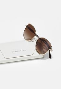 Pruunitooniga kassikuldne päikeseprillid, millel on kuldmetallist raamielemendid, sisaldavad tekstuuriga valget karpi, millel on reljeefne kiri "MICHAEL KORS."