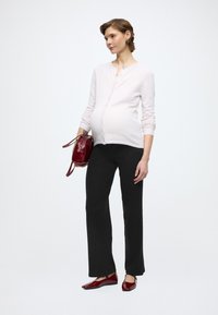 Umstandskleidung bestehend aus einer hellrosa, knöpfbaren Strickjacke, schwarzen, weit geschnittenen Hosen und roten Ballerinas, kombiniert mit einer kleinen roten Tasche.