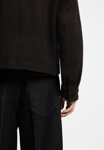 Manche de veste en laine foncée avec poignet boutonné sur un pantalon en denim noir, montrant le bas du dos et la main droite sur un fond clair.