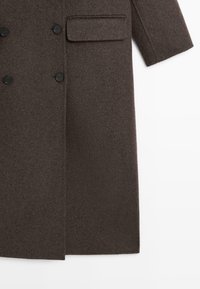 Long manteau marron en mélange de laine avec un design à double boutonnage, deux boutons noirs et une poche poitrine. Tissu texturé avec une finition lisse.