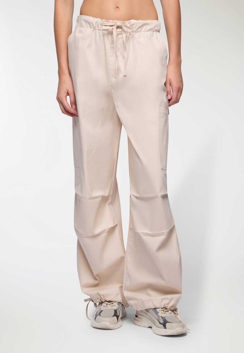 Pantaloni beige con cordoncino in vita, dalla vestibilità ampia e con polsini elasticizzati alle caviglie. Presentano tasche laterali e una superficie liscia. Indossati con sneakers in tonalità neutre.