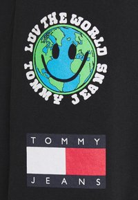 T-shirt en coton noir avec un globe vert et bleu affichant un visage souriant et le texte « AIME LE MONDE », ainsi qu'un logo « TOMMY JEANS » rouge, blanc et bleu.